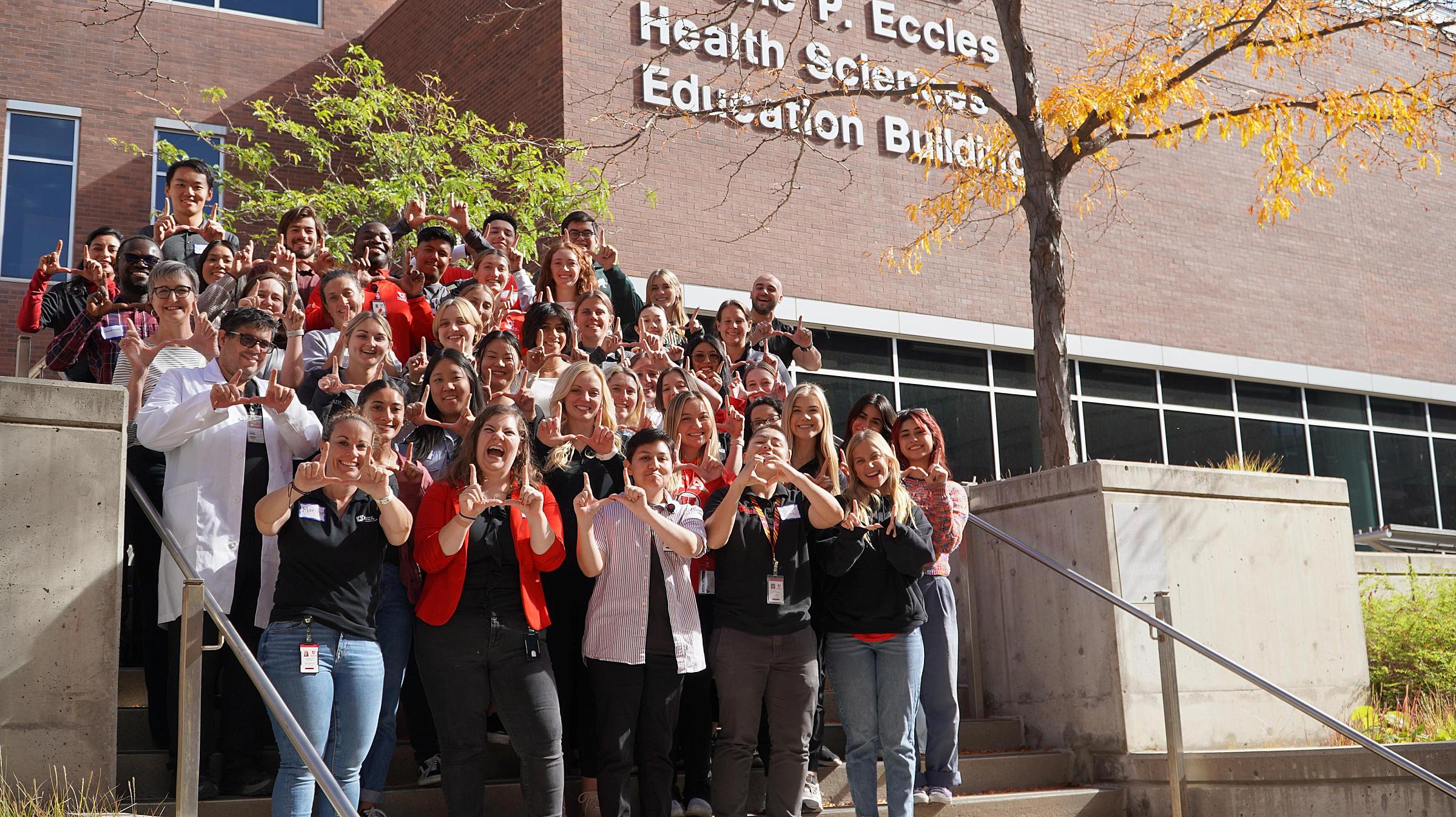 a group of attendees at the October Saturday Academy flash the U outside of the Spencer F. and Cleone P. Eccles Health Sciences Education Building
